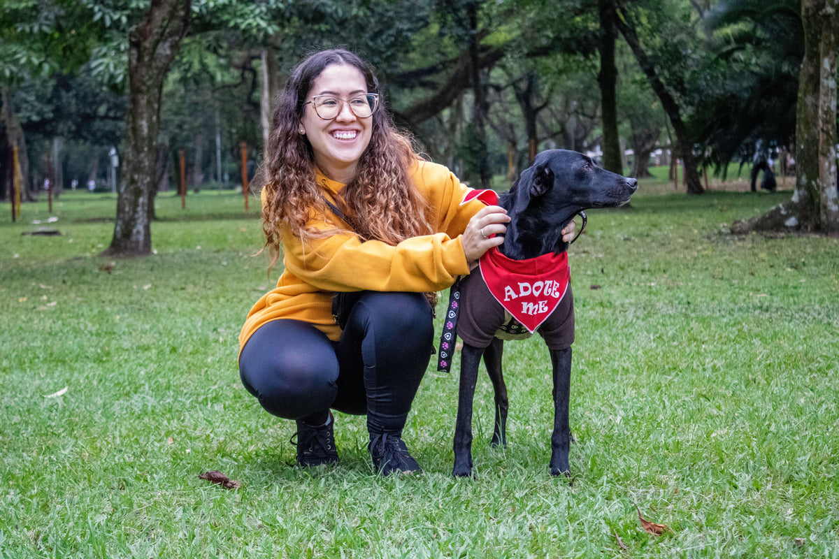 A woman crouching on a grassy park lawn next to a black dog wearing a red bandana that says 'ADOPT ME'.