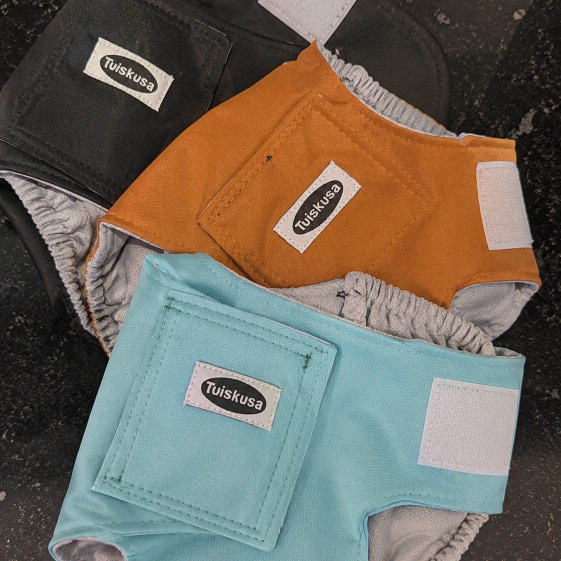 Three dog diapers in black, orange, and blue with 'Tulskusa' branding on a dark surface.