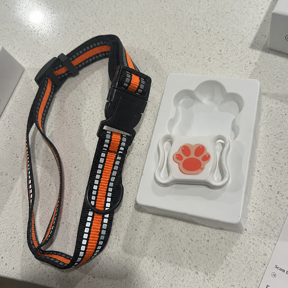 Dog collar with orange and black pattern and a white device with red paw print on a gray surface.