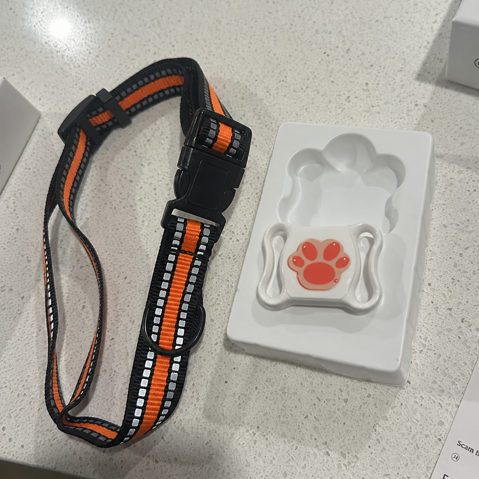 Dog collar with orange and black pattern and a white device with red paw print on a gray surface.