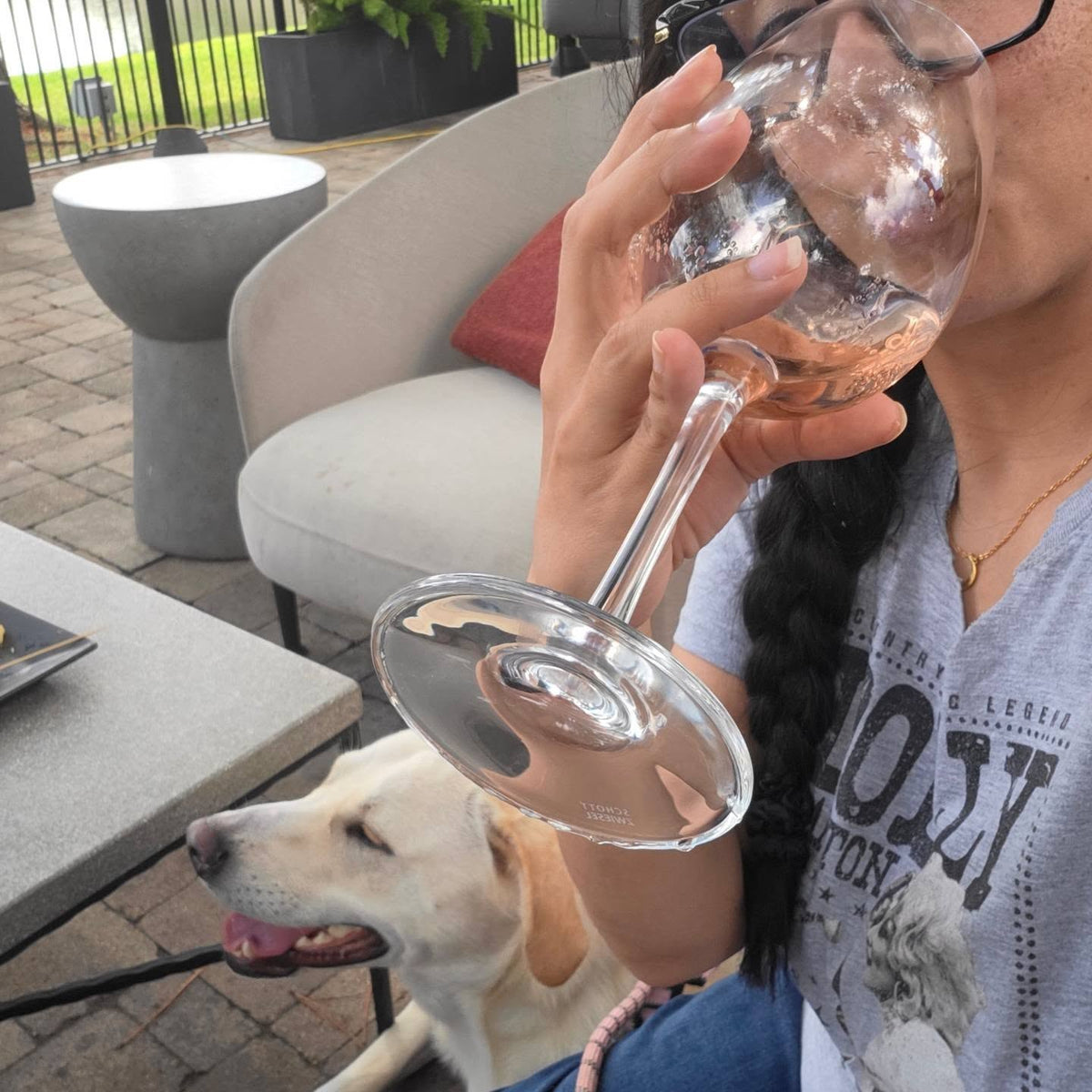 A person holding a clear wine glass with a pink liquid, drinking outdoors on a patio while a light-colored dog sits nearby. The person wears a gray graphic T-shirt and black-framed glasses.