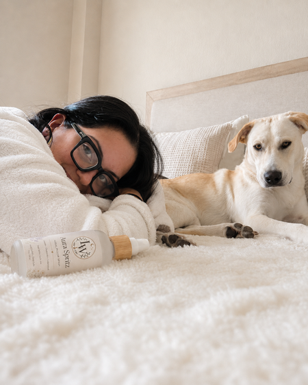 Person lying on a bed with a dog and a bottle of sprayon a pillow